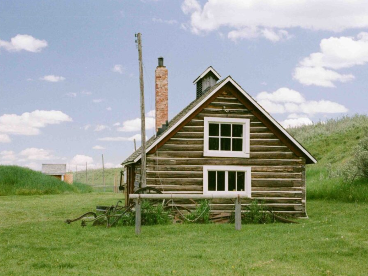 A rustic wooden cabin with a brick chimney stands on a grassy hill under a blue sky with white clouds.