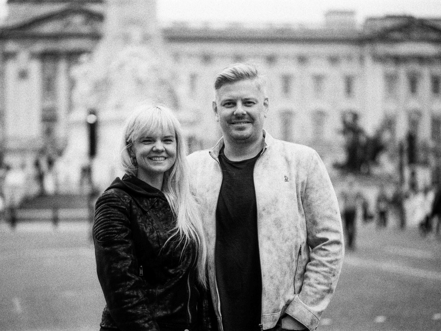 A smiling couple posing together in front of Buckingham Palace on a cloudy day.