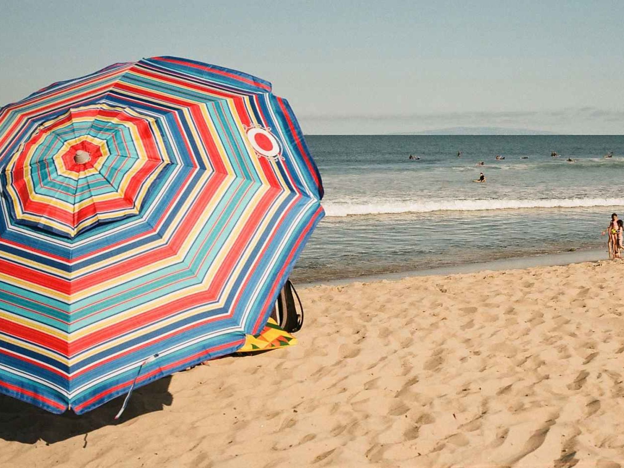Colourful striped beach umbrella on a sandy beach with ocean and people in the background.