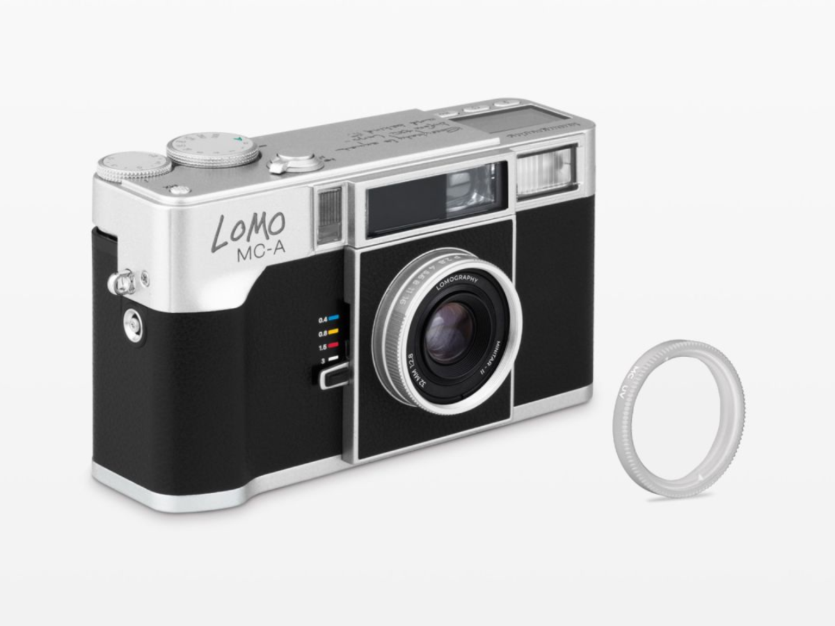 A Lomo MC-A compact camera with a black and silver body, shown alongside a clear circular lens filter on a light gray background.