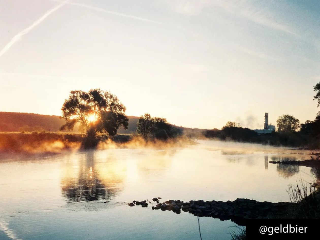 Misty lake at sunrise with trees and a building in the background.