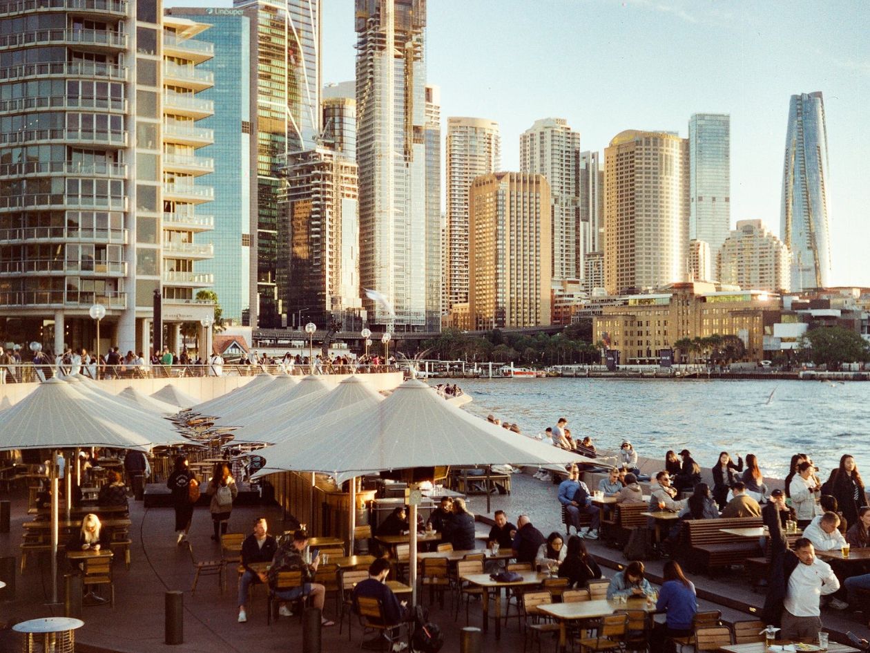 Outdoor dining area with people and umbrellas near a waterfront with tall buildings in the background.