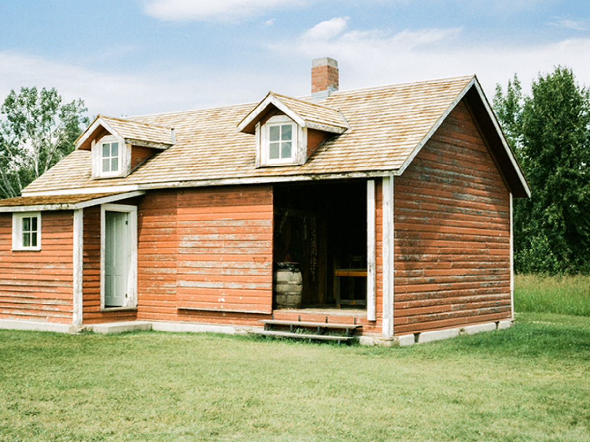 A rustic red-orange wooden cabin with a shingled roof, dormer windows, and an open side door revealing a barrel in the porch, set on a grassy lawn with trees and blue sky in the background.