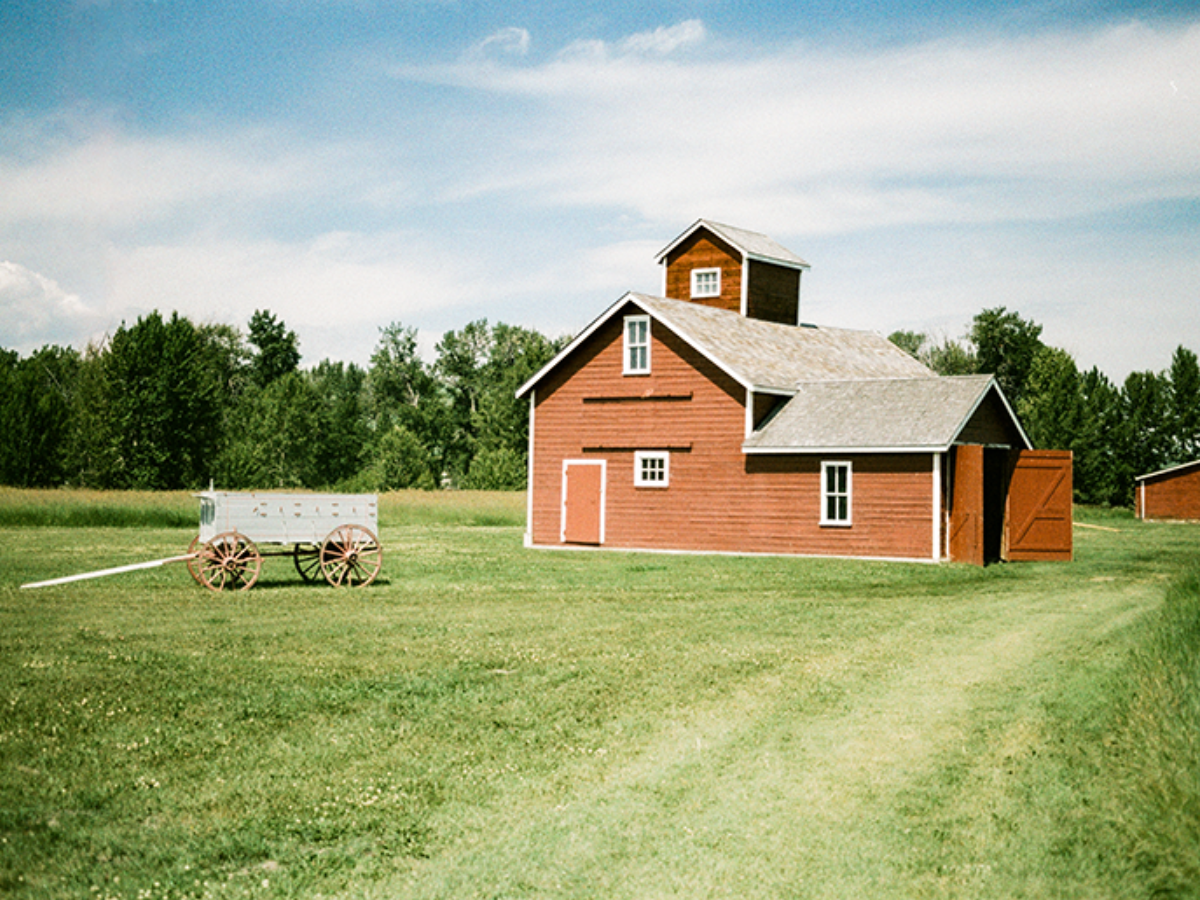 A red wooden barn with a gray shingled roof and white trim stands on a grassy field under a blue sky with clouds. A white wagon is parked to the left, and trees line the background.