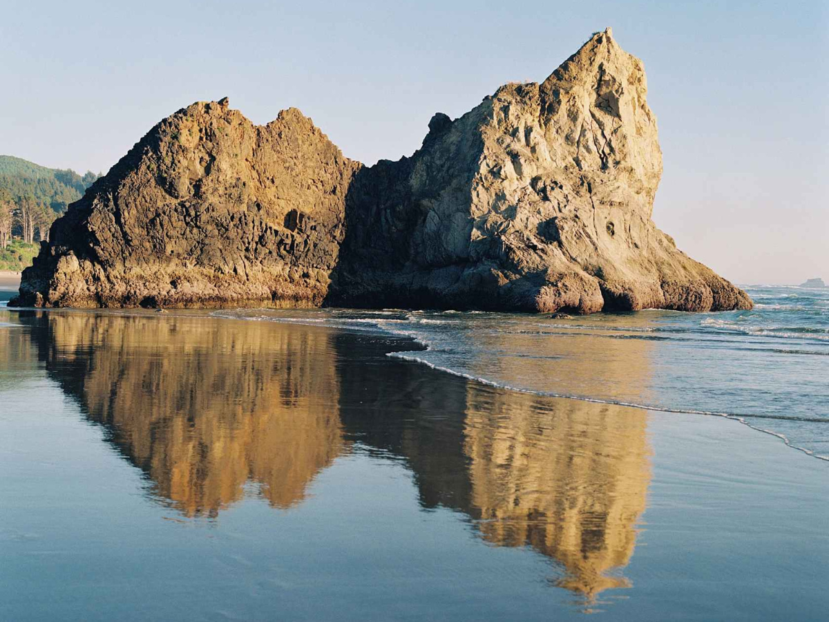 Two large rock formations reflected in calm water on a beach.