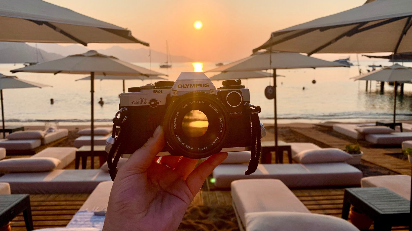 A hand holding an Olympus OM10 film camera at sunrise, with a serene beach, lounge chairs, and umbrellas in the warm glow of the setting sun reflecting on the water.