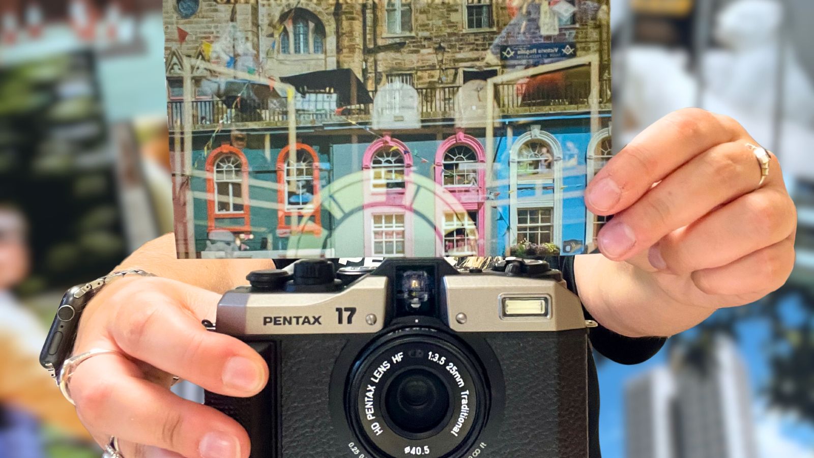 A person holds a Pentax 17 camera with a colorful print of buildings above it, against a blurred background of other vibrant images.
