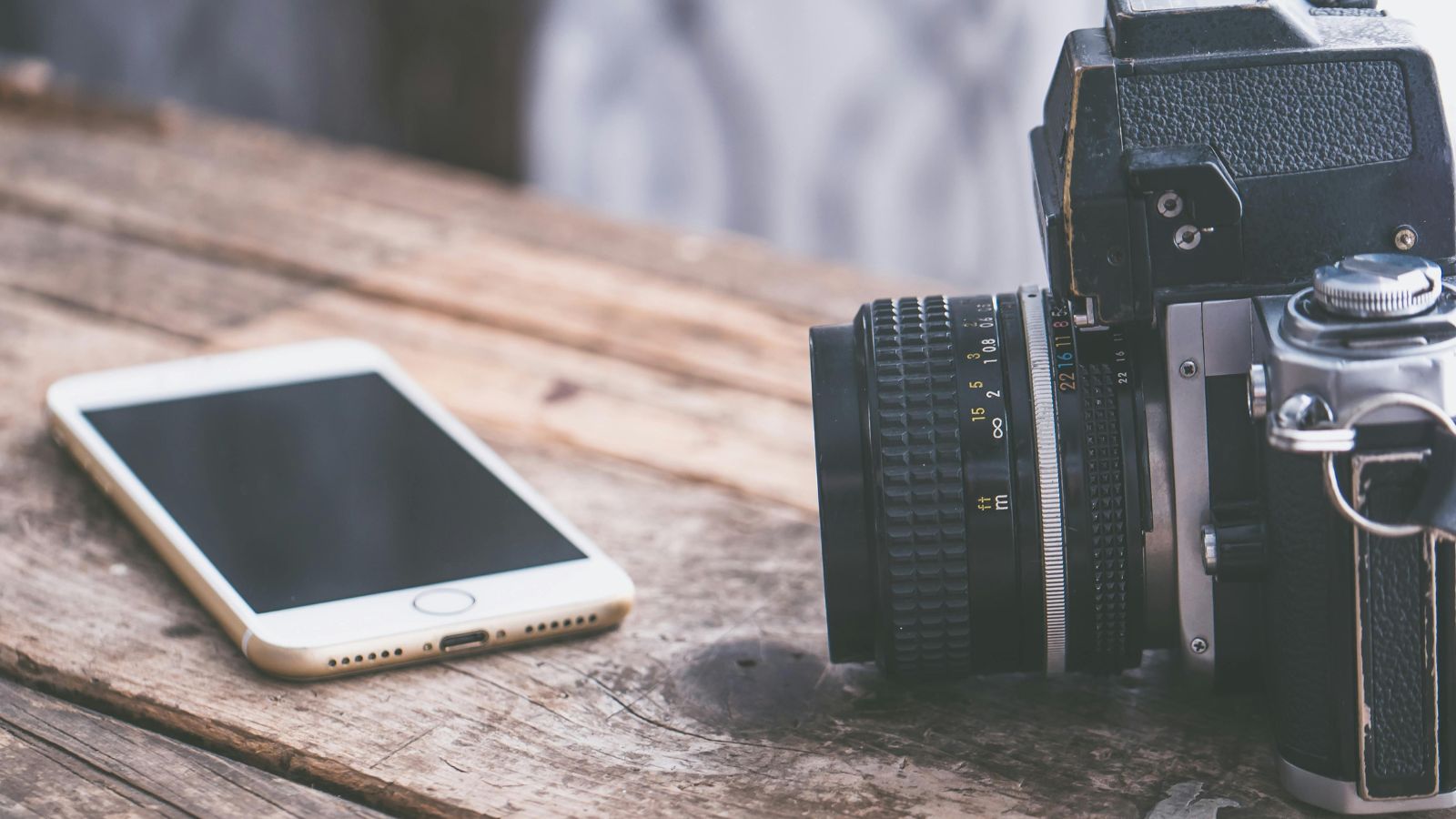 A smartphone and a vintage film camera sit side by side on a wooden table.