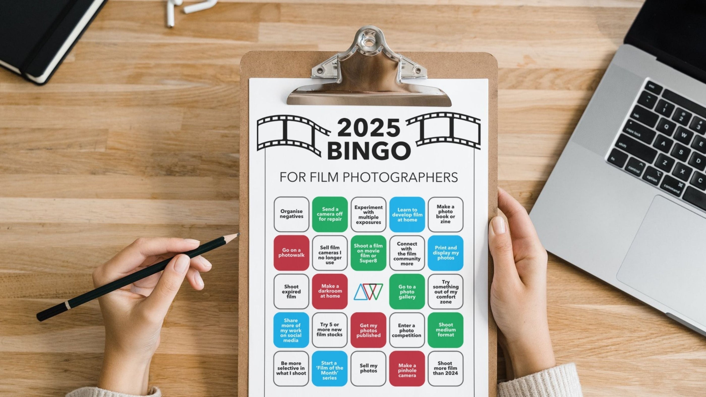 A clipboard with a "2025 Bingo for Film Photographers" card sits on a wooden desk alongside a laptop, notebook, and pencil.