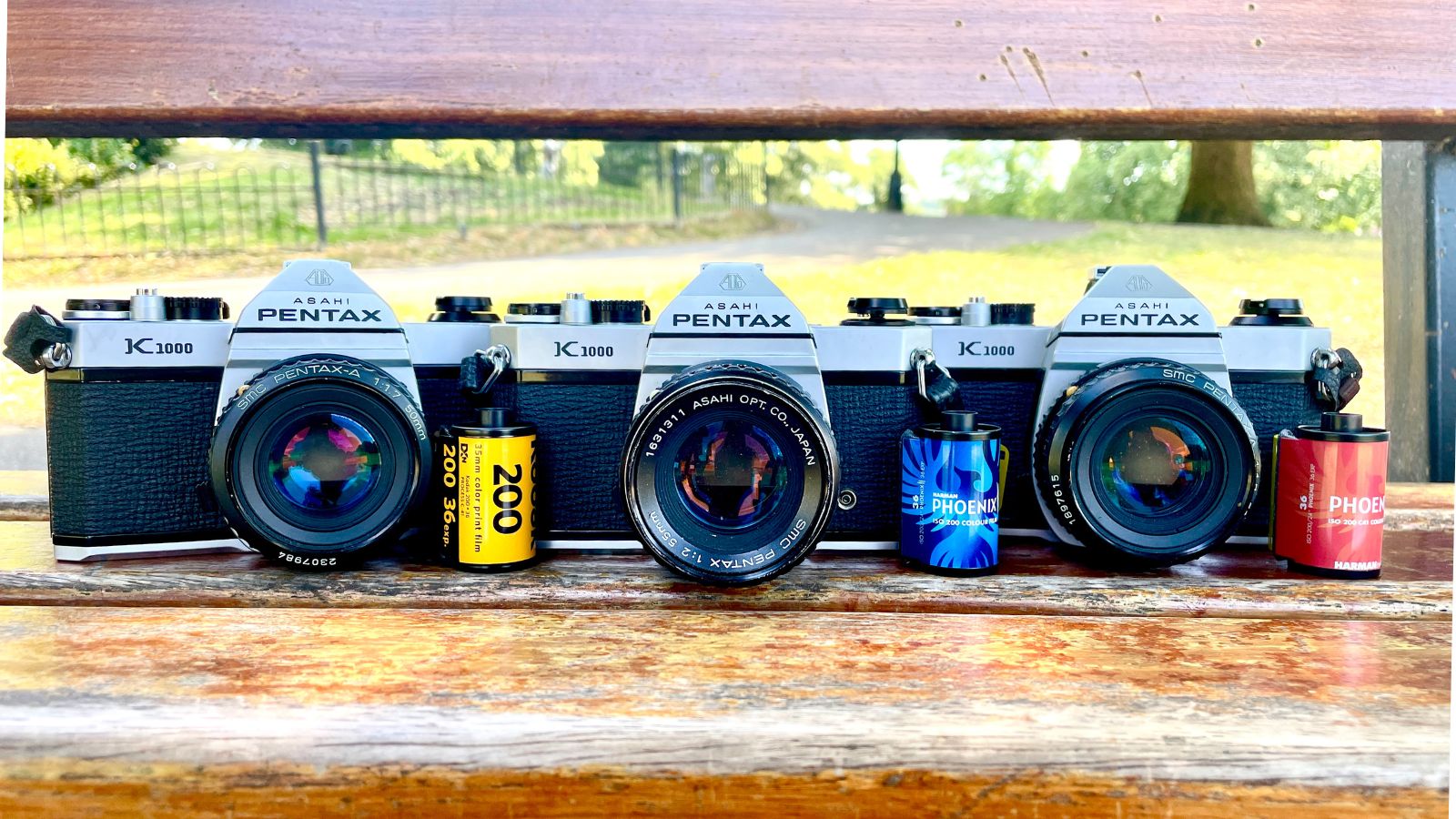 Three Pentax K1000 film cameras on a bench with Kodak Gold, Harman Phoenix, and Phoenix II ISO 200 film rolls displayed in front.