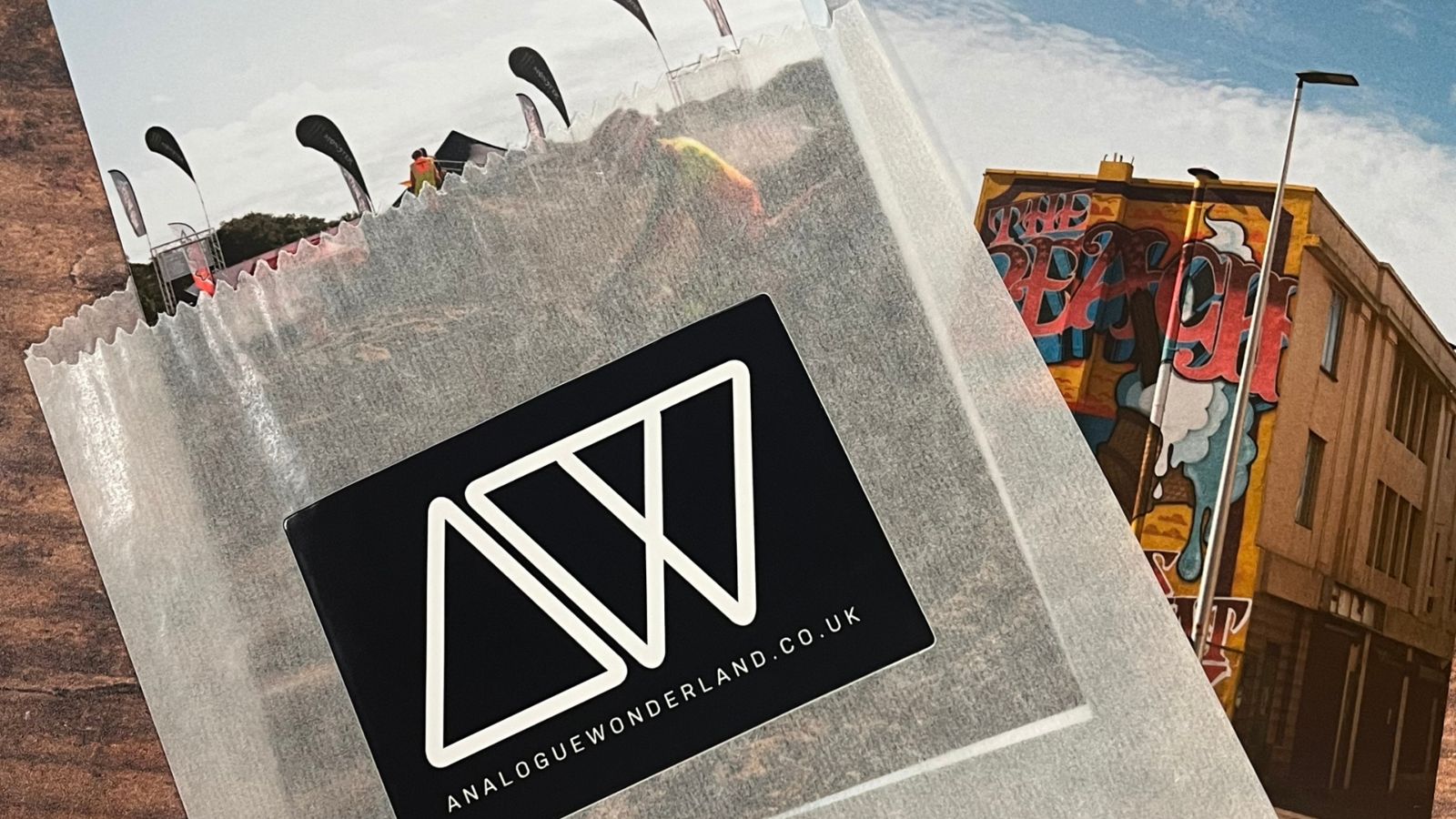 A close-up of an Analogue Wonderland logo sticker on a translucent paper bag, with one print inside and one a wooden table featuring a vibrant mural and blue sky.