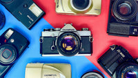 A flat lay of various vintage film cameras arranged on a red and blue split background.