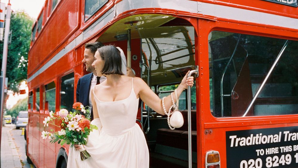 Bride and groom stepping off a vintage red London bus, holding flowers and smiling on their wedding day.