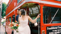 Bride and groom stepping off a vintage red London bus, holding flowers and smiling on their wedding day.
