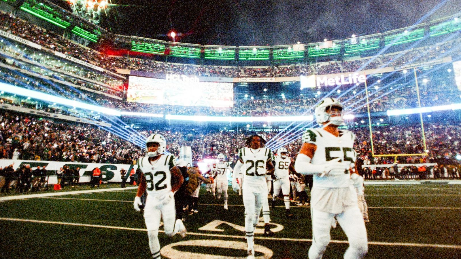 New York Jets players enter the field at MetLife Stadium, with bright stadium lights and a full crowd in the background, captured with a vibrant film aesthetic