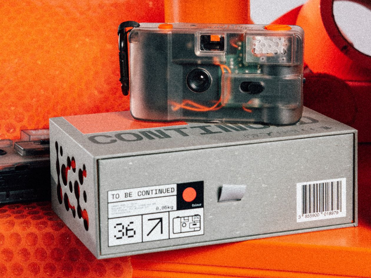 Transparent 35mm film camera labelled "TO BE CONTINUED" on a grey box with icons and a barcode, against an orange background.