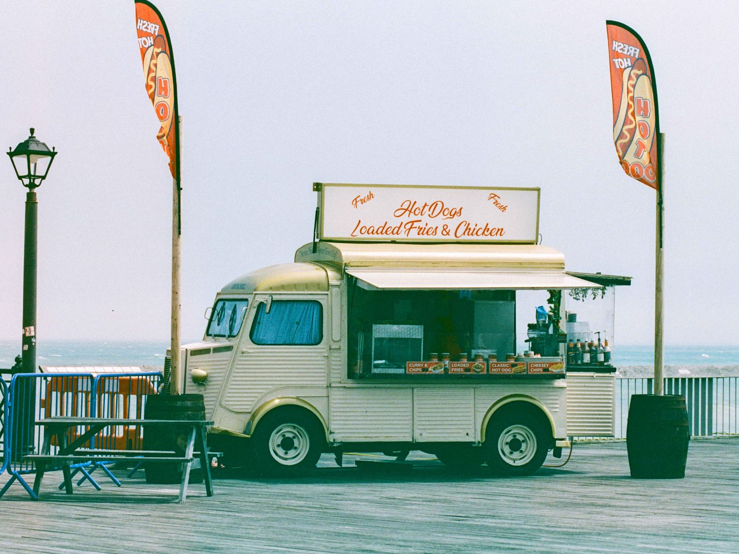 A vintage-style food truck on a seaside boardwalk selling hot dogs, loaded fries, and chicken.