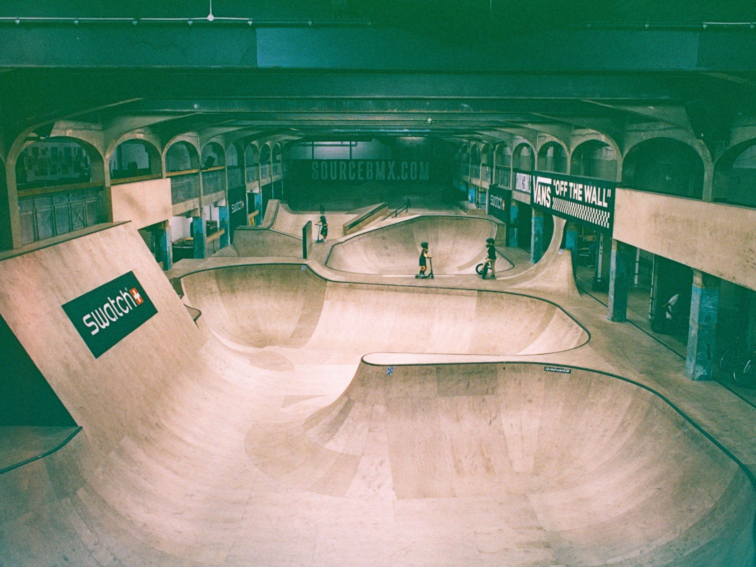 A large indoor skatepark with wooden ramps and BMX riders, featuring Swatch and Vans branding.