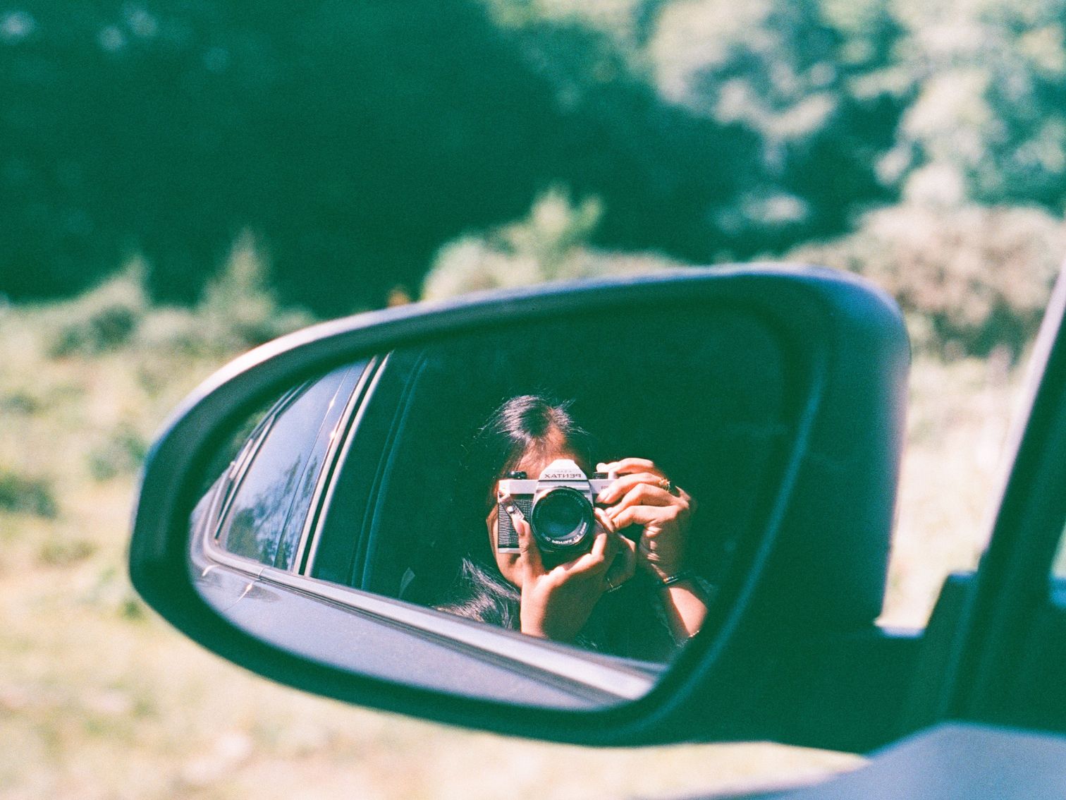 A woman photographing through a car’s side mirror using a Pentax film camera, with greenery in the background.