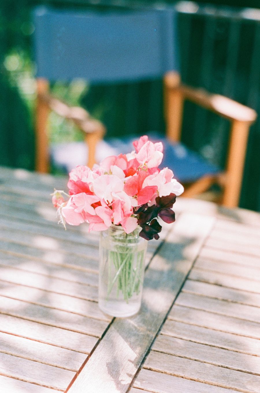 A small glass vase with a bunch of pink and red sweet peas on a sun-dappled wooden table.