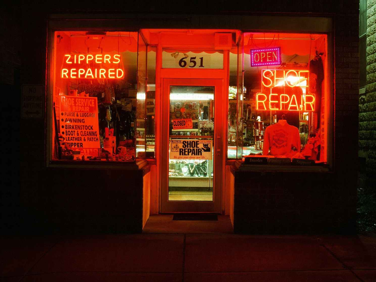 A storefront at night with bright red neon signs reading 'ZIPPERS REpaired' and 'SHOE REPAIR' around a central door marked '651'.