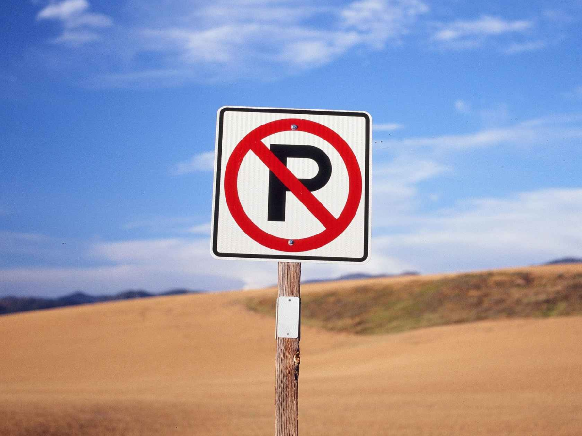 A square white 'No Parking' sign with a red circle and slash over a black letter P, mounted on a wooden post in an outdoor desert landscape under a blue sky.