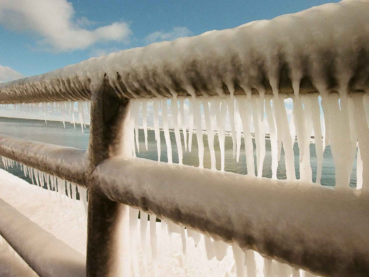Close-up of a metal railing covered in thick icicles, with snow and a frozen body of water visible in the background under a blue sky.
