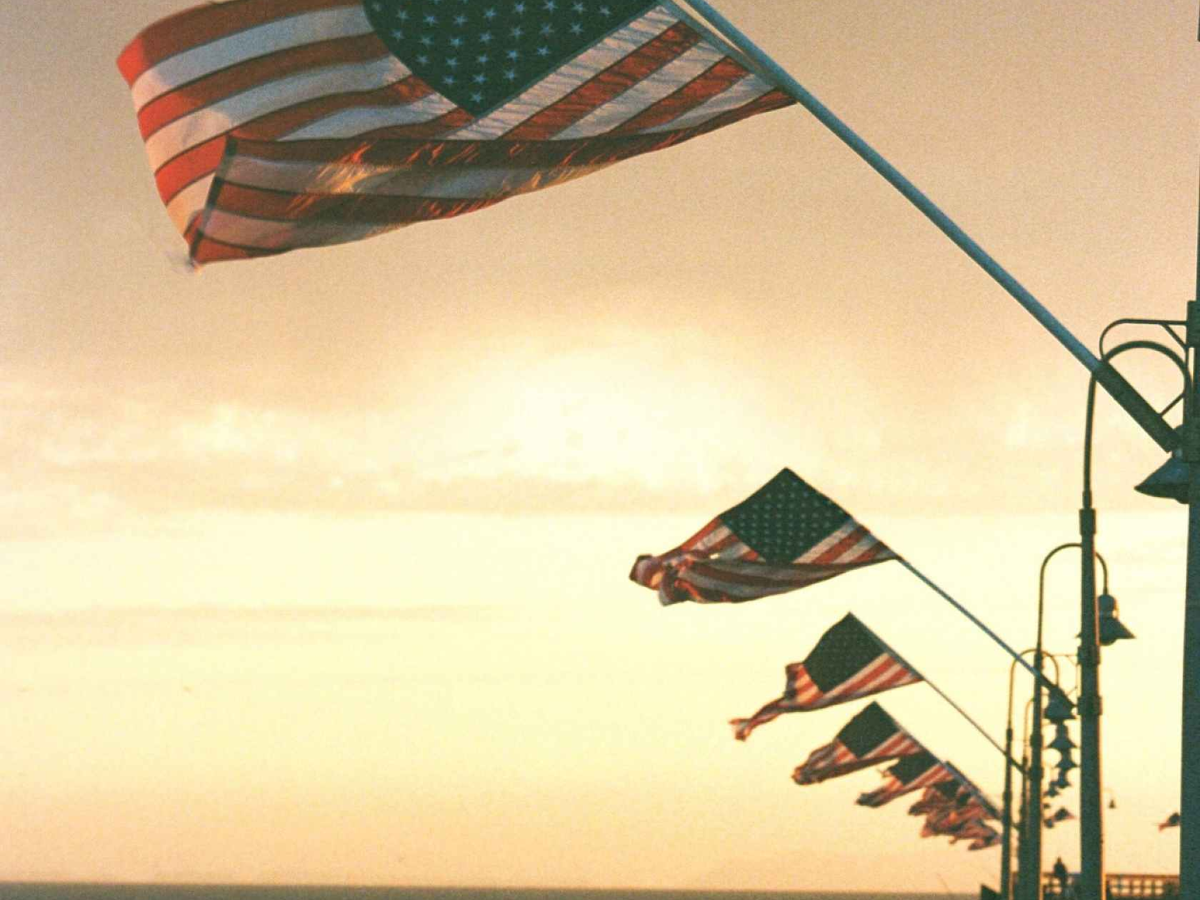 Multiple American flags waving along a waterfront at sunset, with a bright sunlit sky and pole lines receding into the distance.