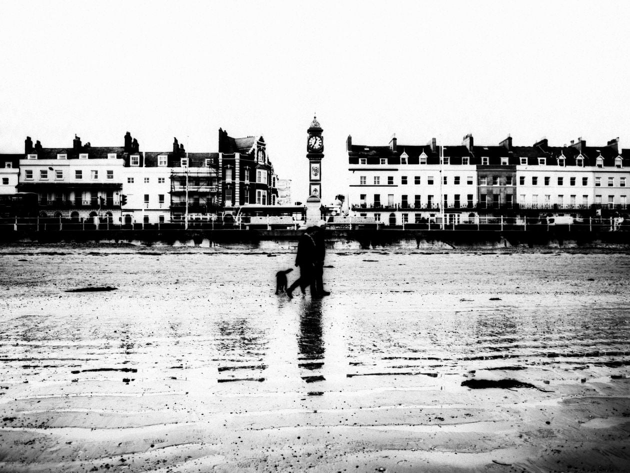 Black and white photo of a person walking a dog on a beach with buildings in the background.