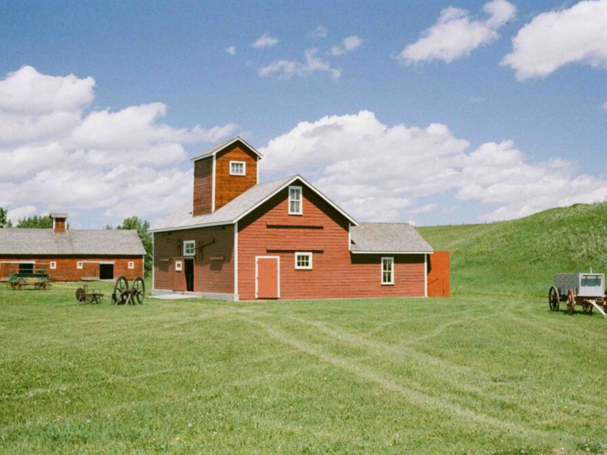 A red wooden barn with a tall smokestack and gray roof, situated on a grassy hill under a blue sky with white clouds. A smaller matching barn is visible to the left, with a horse-drawn cart on the right.