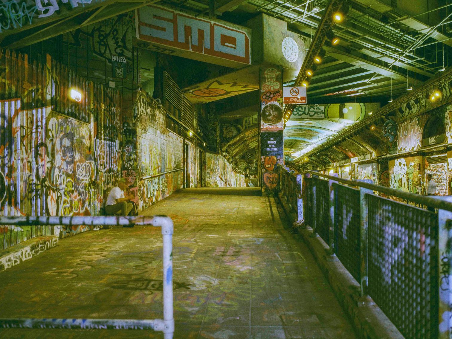 A man sits alone in a graffiti-covered tunnel beneath a bridge, illuminated by moody yellow lights in a richly textured urban landscape. Captured by Heather Hughes using a FujiGSW690ii film camera loaded with Harman Phoenix II 120 film.