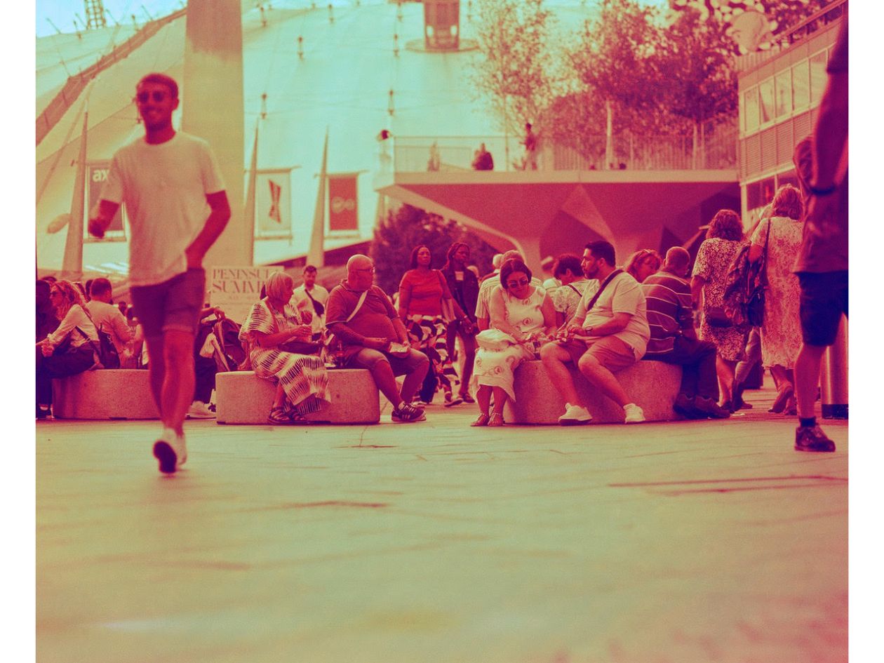 A busy outdoor scene with people sitting and walking, bathed in warm red and orange tones.