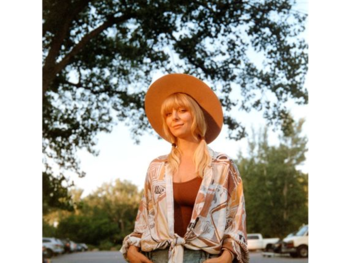 A woman wearing a wide-brimmed tan hat, a brown tank top, and a patterned open-front shirt over light blue jeans, standing outdoors under trees.
