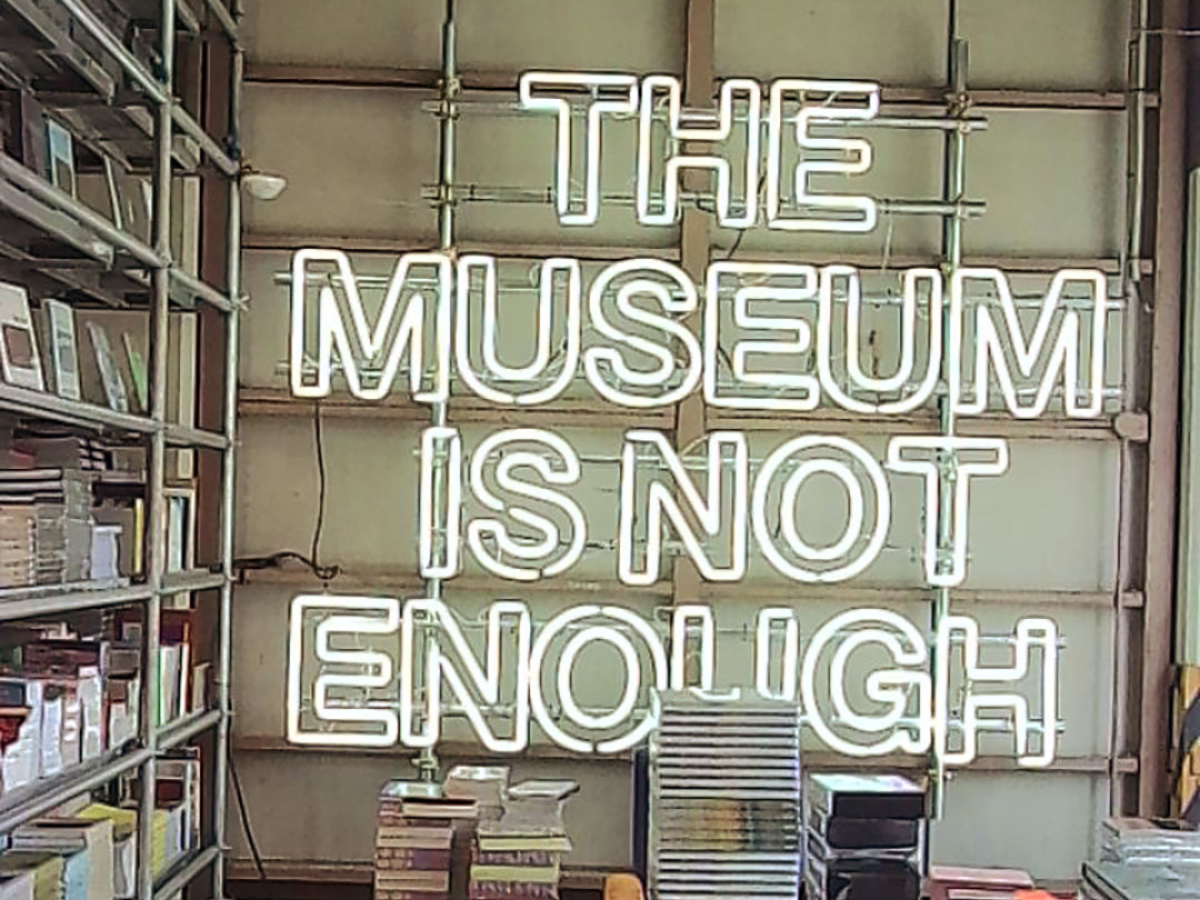 Sample Image | Neon sign in a room with bookshelves and books, reading 'THE MUSEUM IS NOT ENOUGH'.