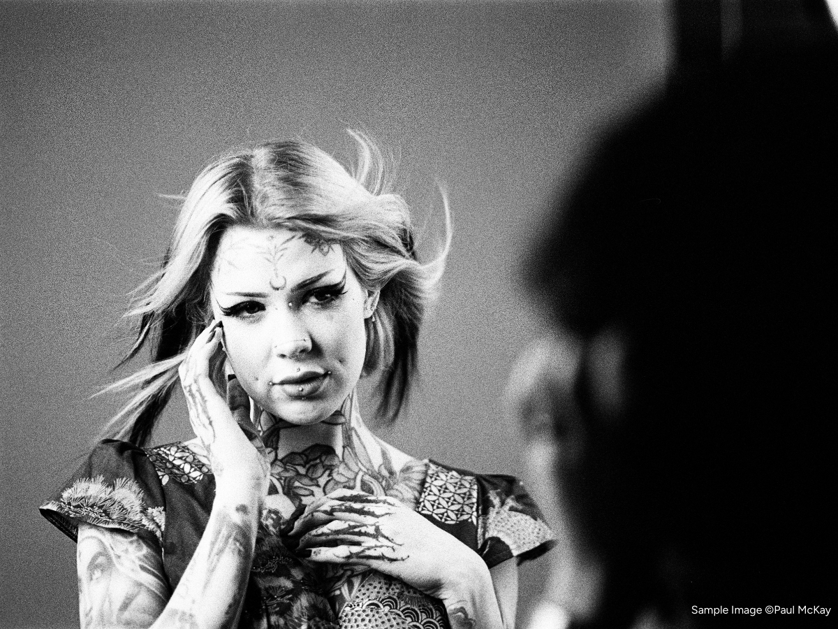 Black and white photo of a person with tattoos and patterns on clothing, holding their hand to their face.