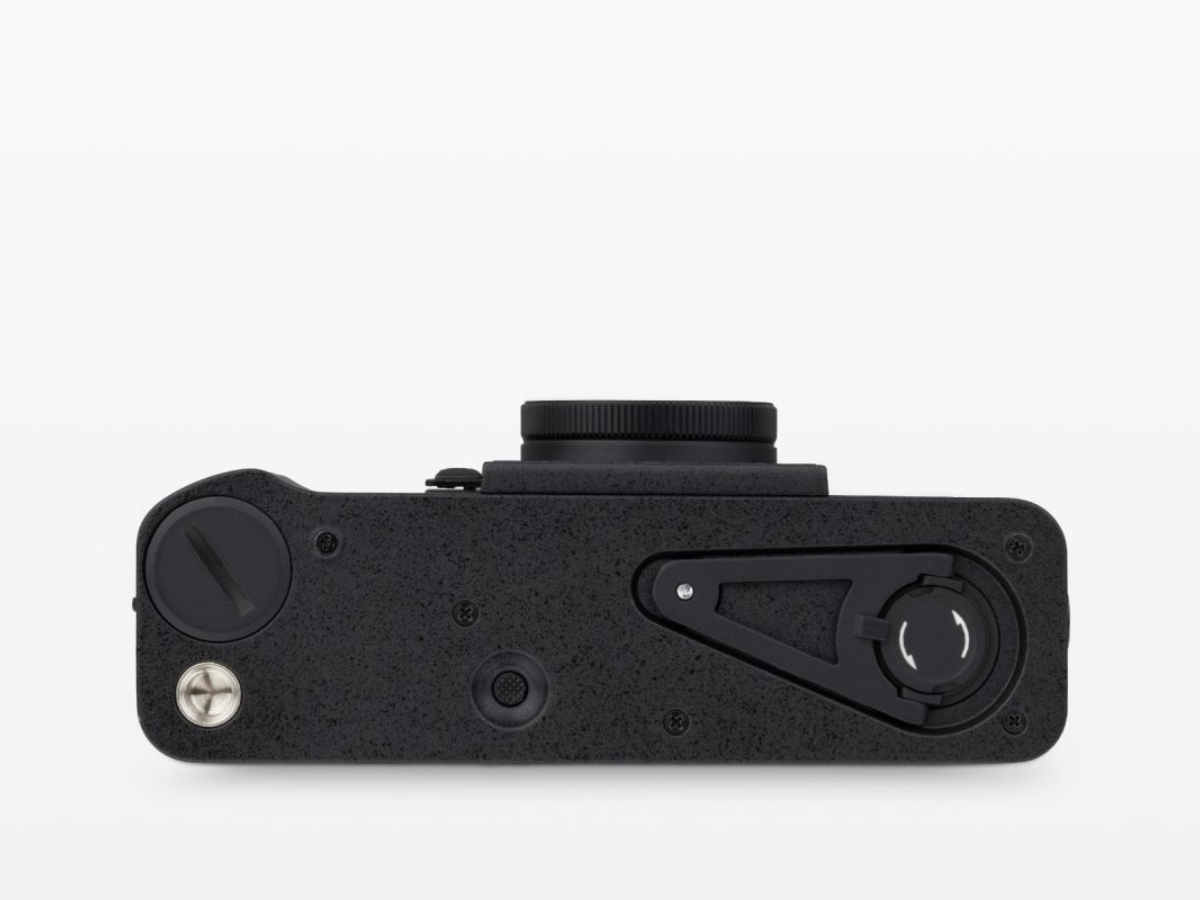 A close-up top-down view of a black textured camera body, showing the lens mount, control dials, and a silver screw on a clean white background.