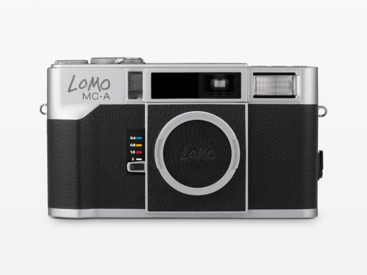A Lomo MC-A compact film camera with a silver metal body and black textured leatherette grip, shown from the front on a light gray background.
