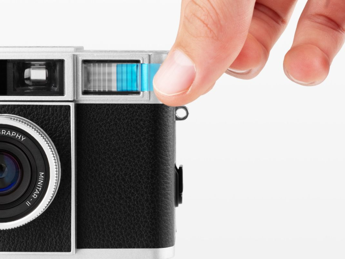 A close-up of a camera body with a hand pressing a blue release button on the top right, shown against a clean white background.