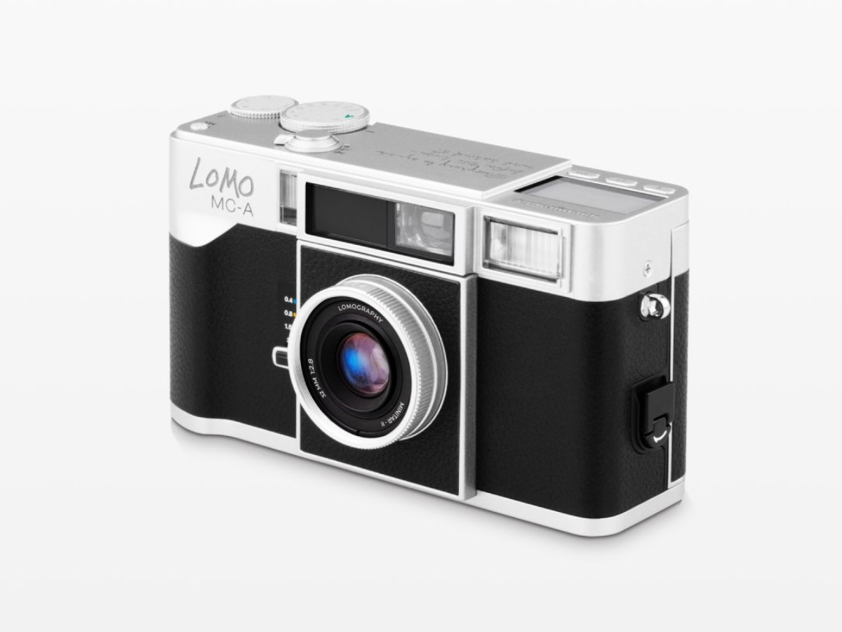 A Lomo MC-A compact camera with a black textured body and silver metal accents, shown at a three-quarter angle on a light gray background.