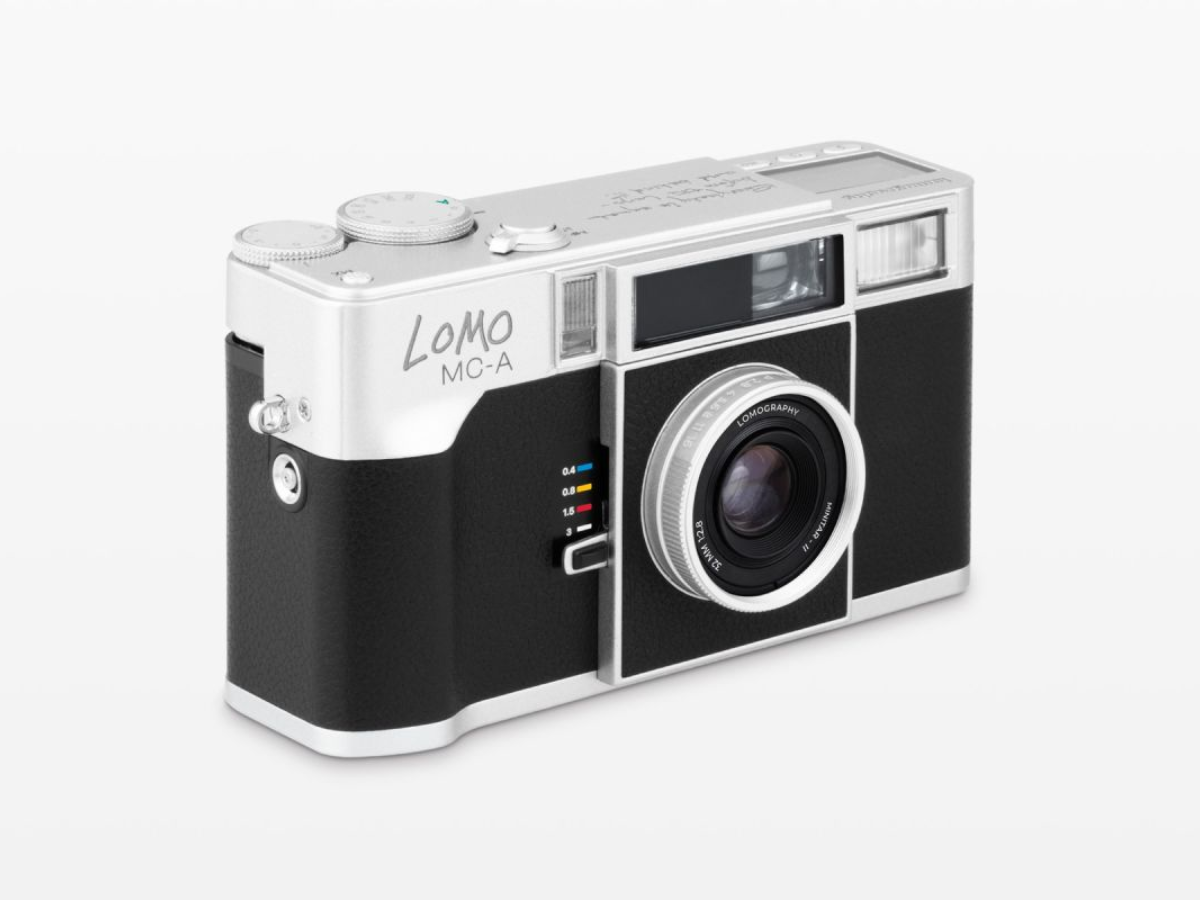 A Lomo MC-A compact camera with a black and silver body, shown at a three-quarter angle on a light gray background.
