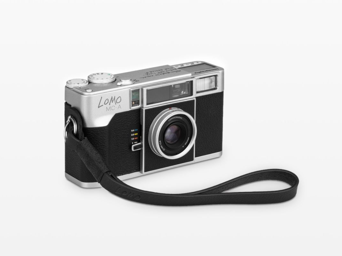 A vintage Lomo MC-A rangefinder camera in silver and black, featuring a black wrist strap, photographed on a clean white background.