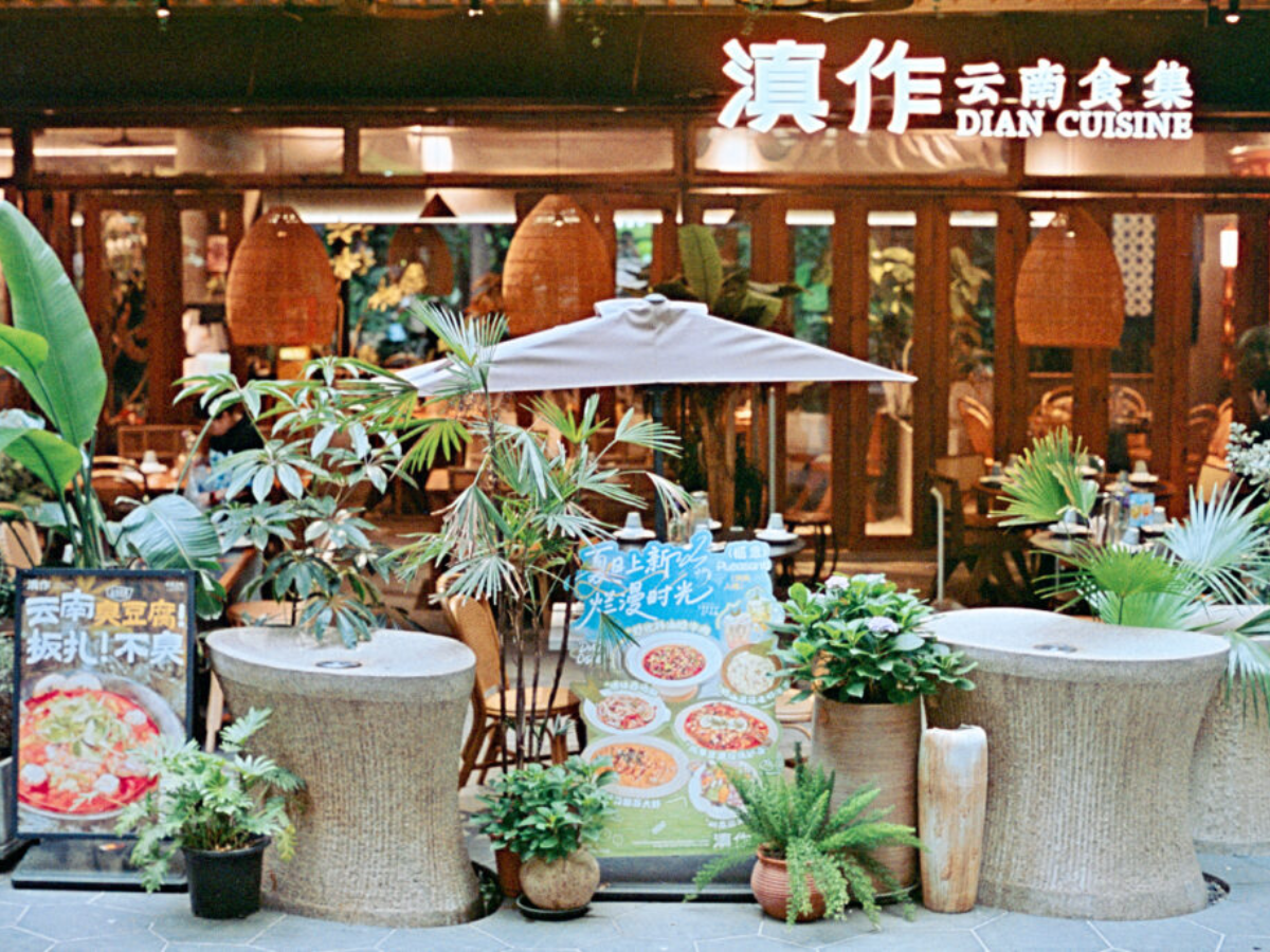 Outdoor dining area of Dian Cuisine restaurant with woven cylindrical tables, potted plants, a large umbrella, and a restaurant interior with wooden framing and Chinese signage in the background.