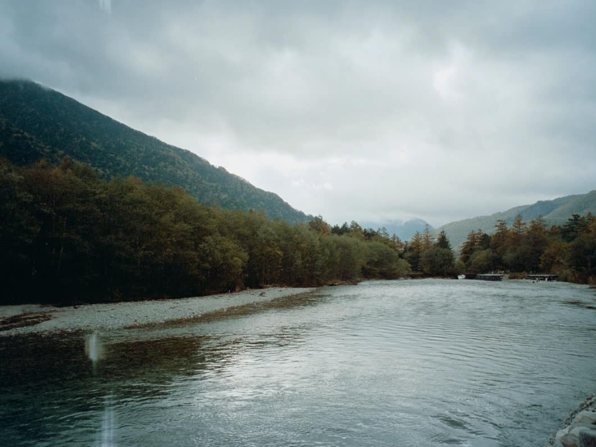 River flowing through a forested area with mountains in the background on a cloudy day. (c) Sample Photos by @notmegapixels