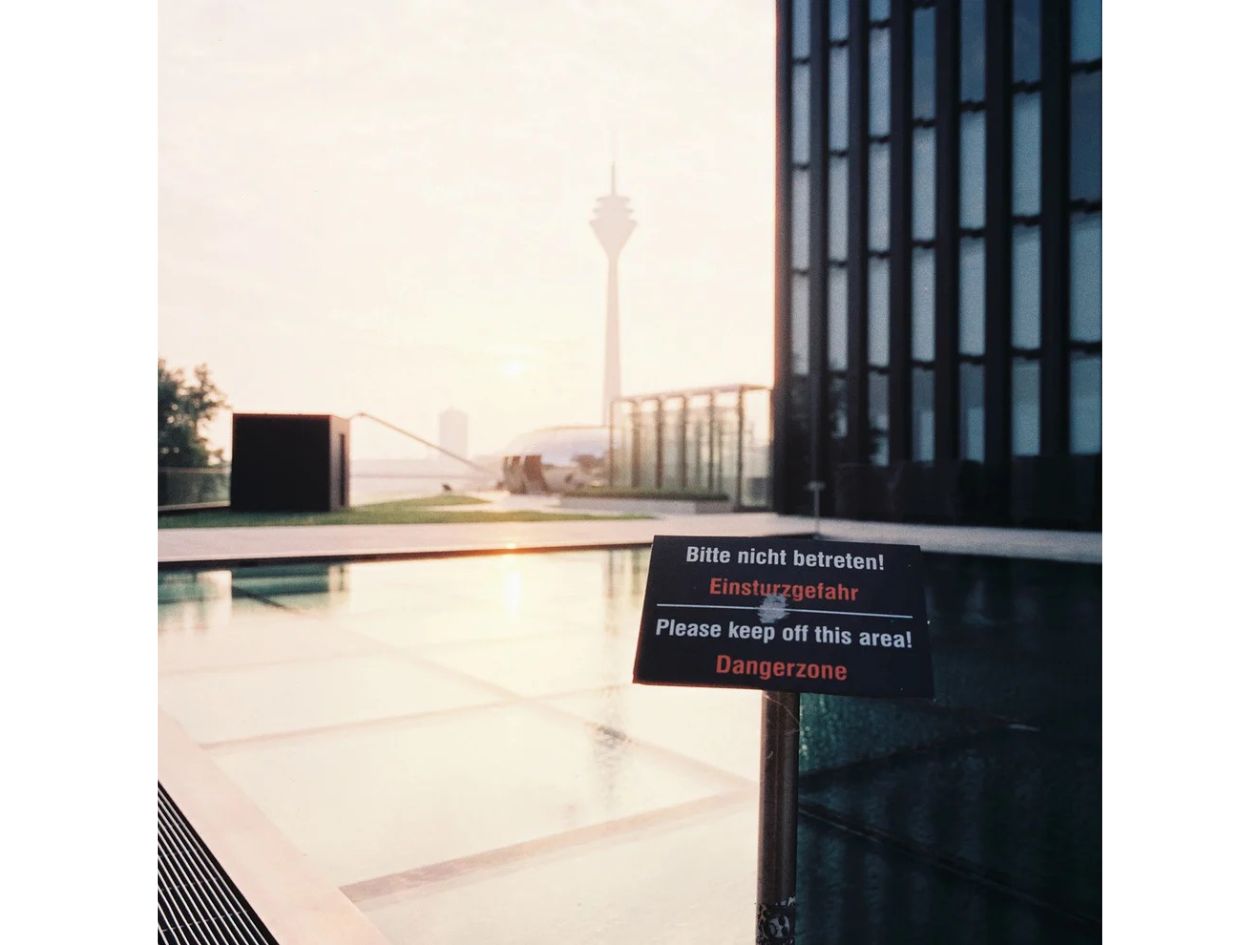 Sign warning 'Please keep off this area' in front of a modern building with a tower in the background.