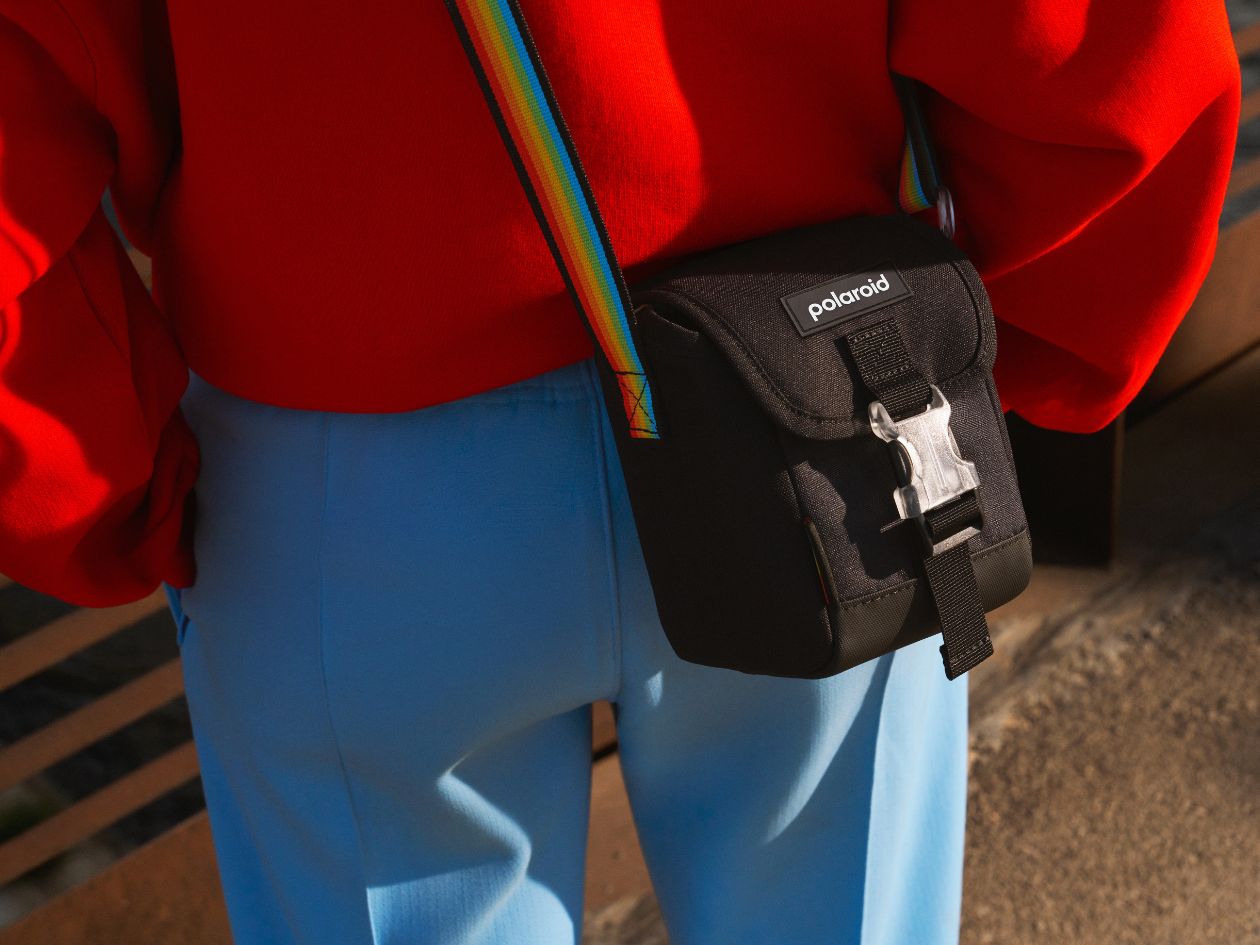 Person wearing a red jacket and blue pants with a black Polaroid camera bag.