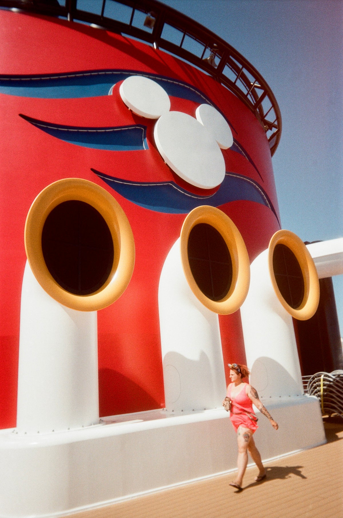 Sample photo for the Kodak EC35 film camera. A woman in a pink swimsuit walks along the deck of a cruise ship, with the iconic red and white Disney funnel and Mickey Mouse silhouette in the background under a clear blue sky.