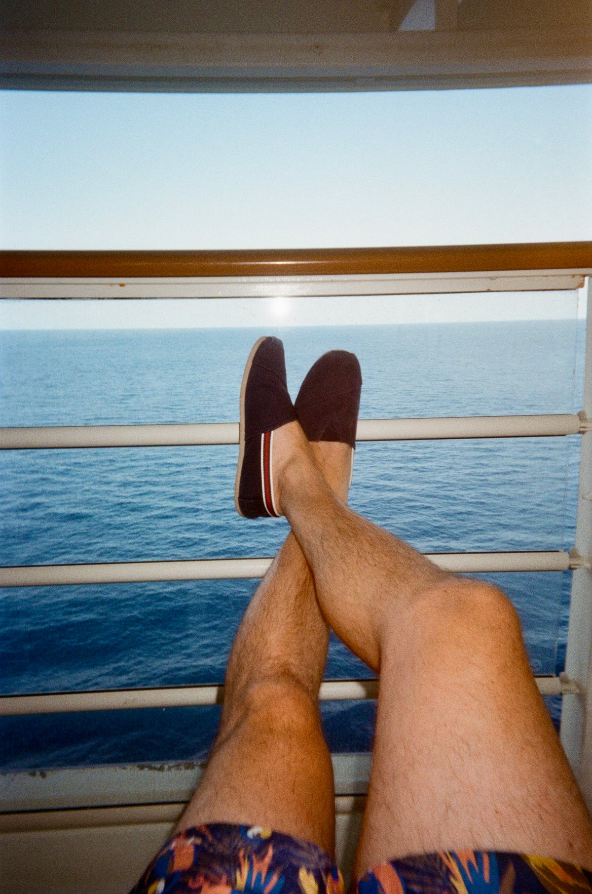 Sample photo for the Kodak EC35 film camera. A person's legs crossed with feet resting on a balcony railing overlooking the blue ocean, wearing dark slip-on shoes and colorful patterned swim trunks.