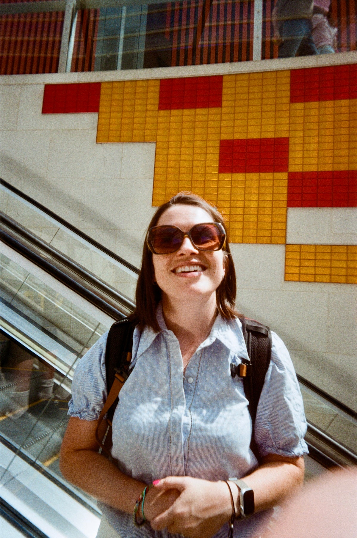 Sample photo for the Kodak EC35 film camera. A woman wearing sunglasses, a light blue patterned button-up shirt, and a black backpack stands on an escalator in front of a yellow and red tiled wall.