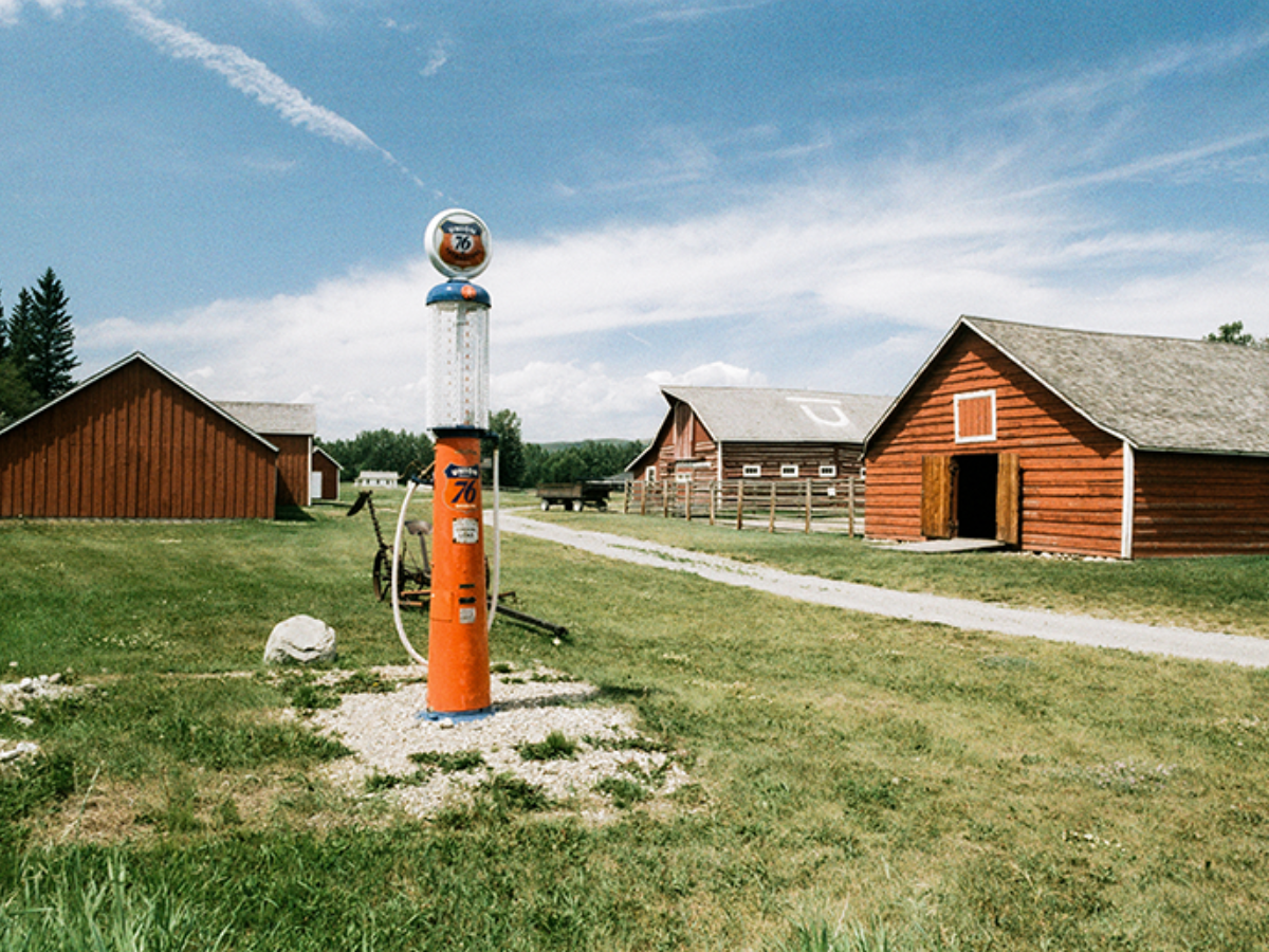 A vintage orange and white gas pump stands on a gravel patch in a grassy field, with red-brown barns and a dirt road under a blue sky in the background.