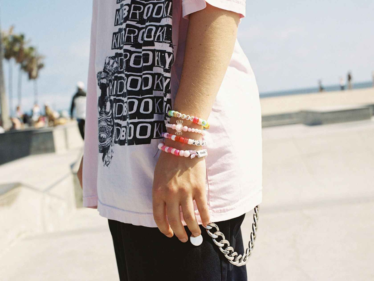 Person wearing a white t-shirt with black text and graphics, holding colorful bracelets on a blurred outdoor background.